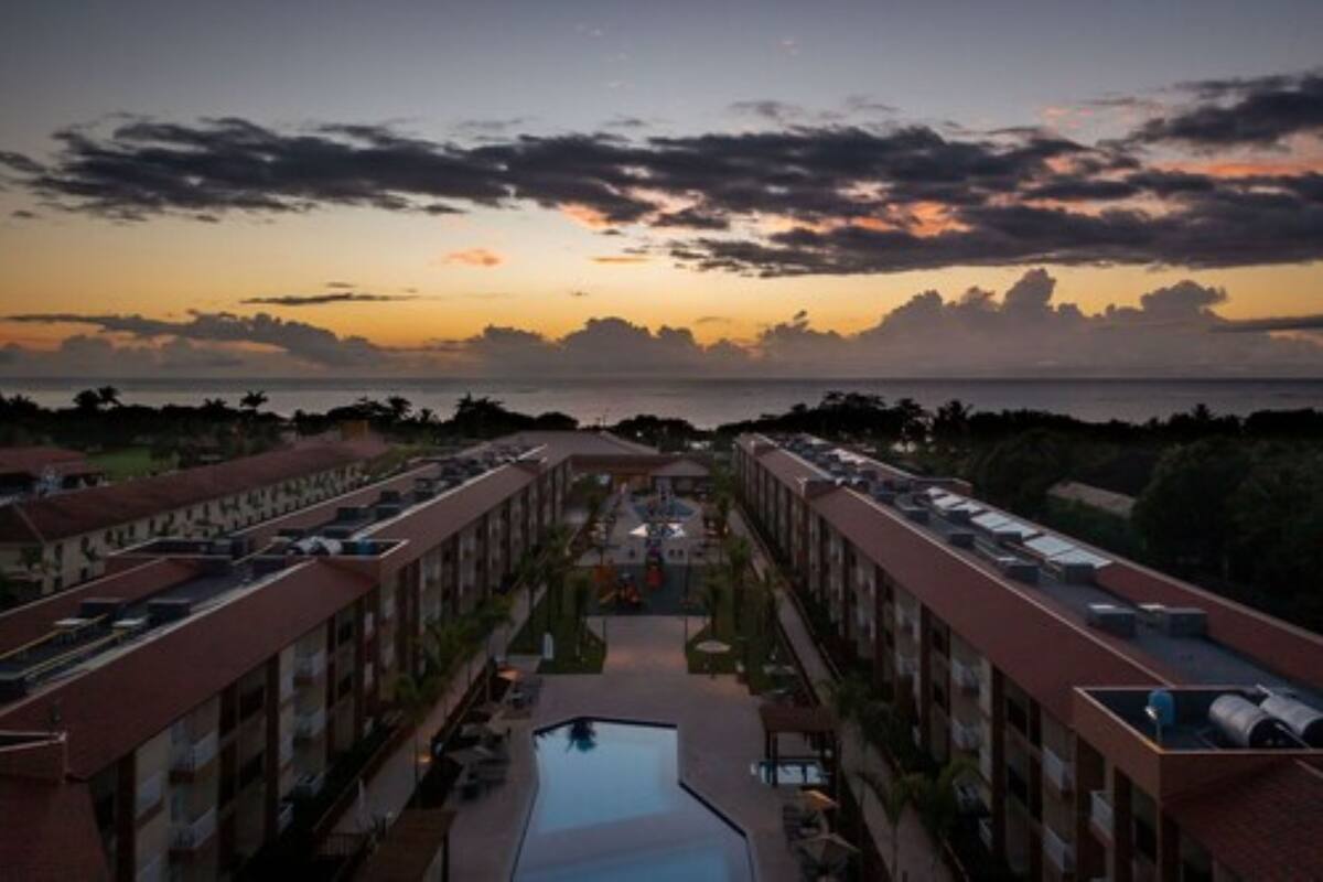 A hotel with a central pool, multiple floors, and a view of the sea at sunset.