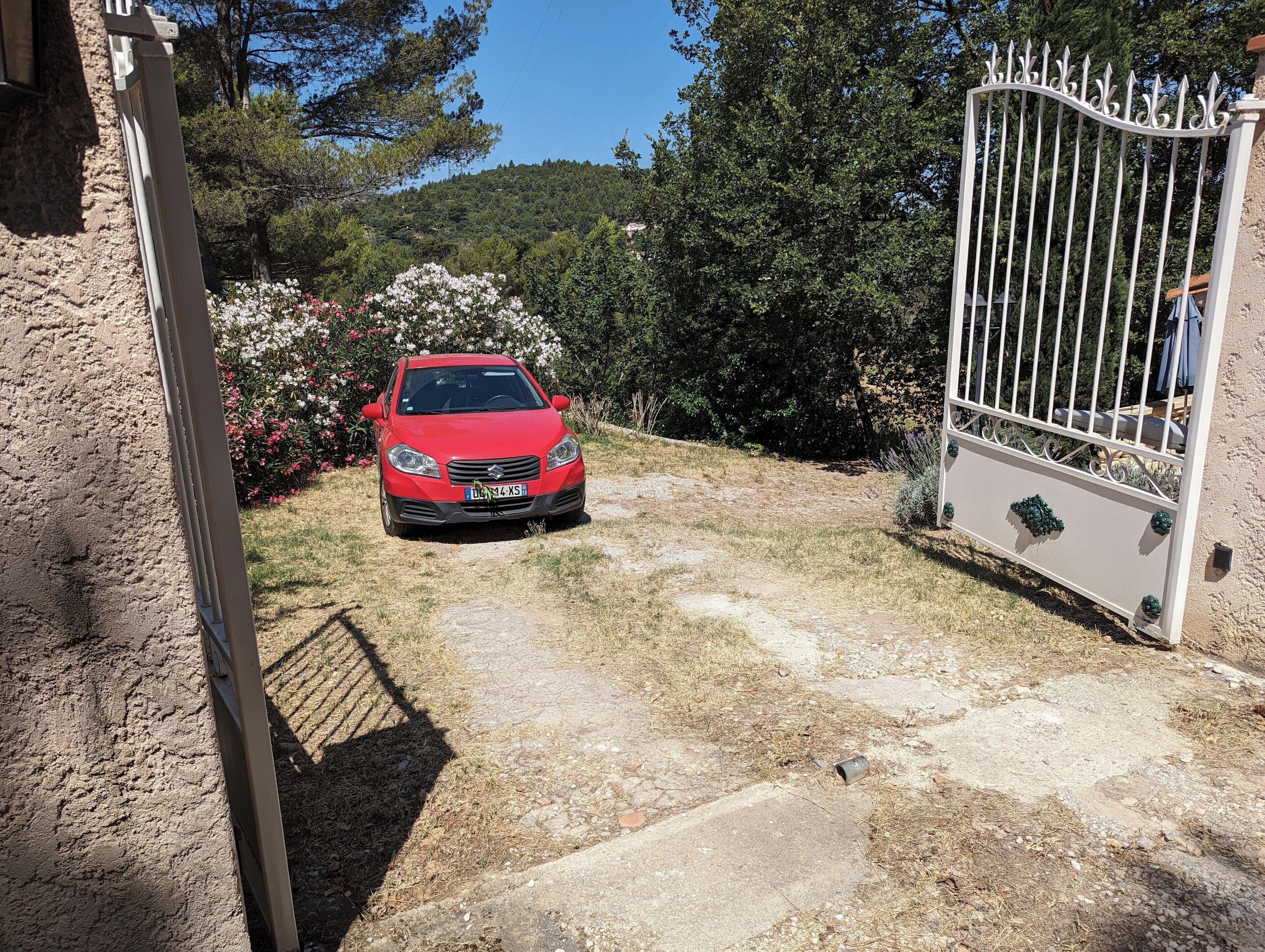 A red car parked in a driveway with a gated entrance, surrounded by greenery and a hill in the background.