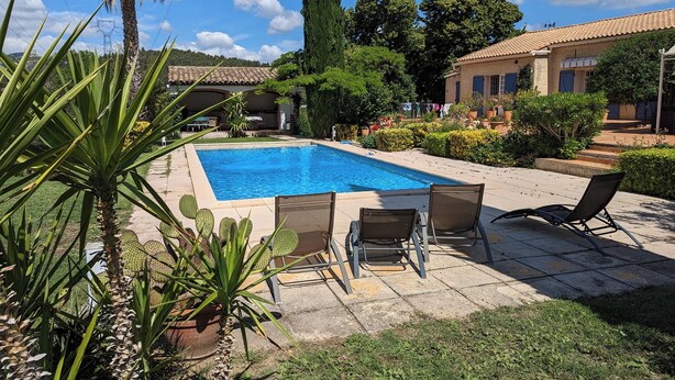 Une piscine rectangulaire, bordée d’une terrasse carrelée, entourée de verdure et agrémentée de quelques chaises.