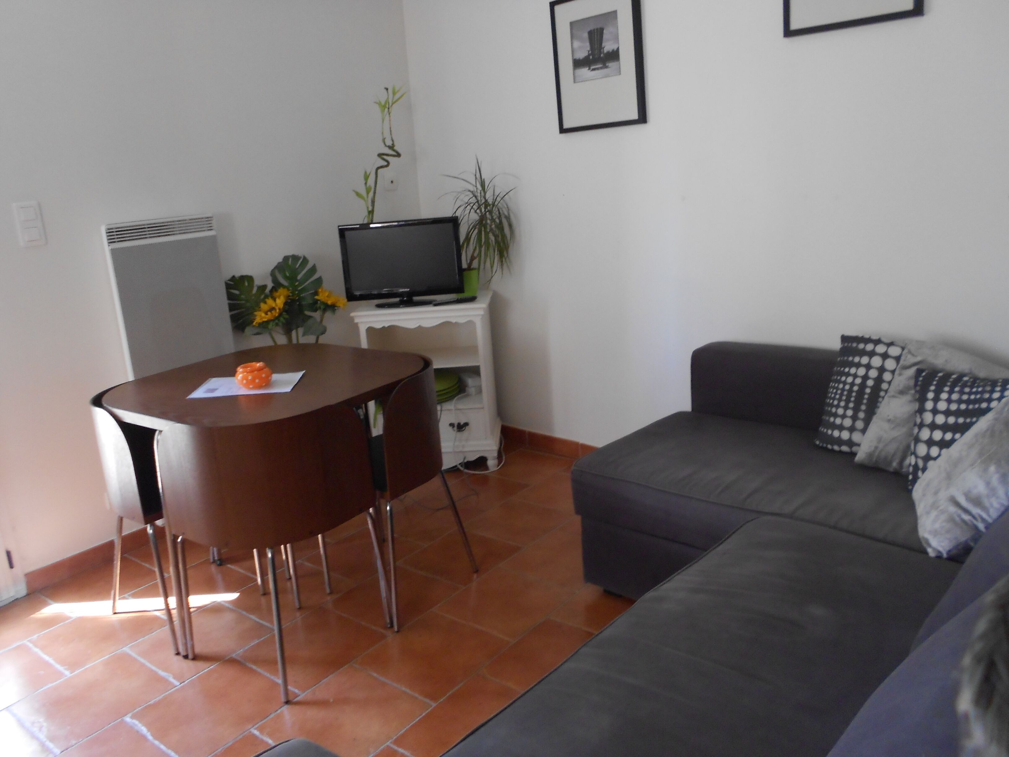 A living room with a grey sofa, a small round table with two chairs, a TV on a white stand, and a framed picture on the wall.