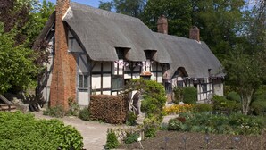 Une maison traditionnelle à colombages, avec un toit de chaume, des cheminées en briques et un jardin où poussent diverses plantes et fleurs.