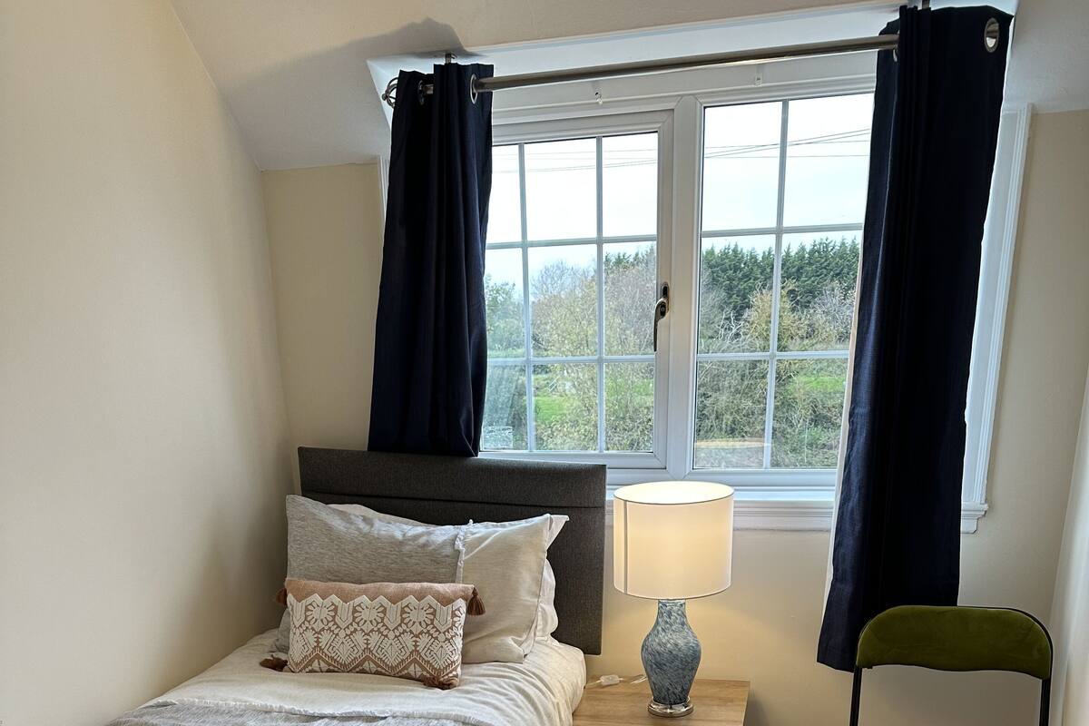 A bedroom with a bed, a nightstand, a lamp, a chair, and a window with curtains.