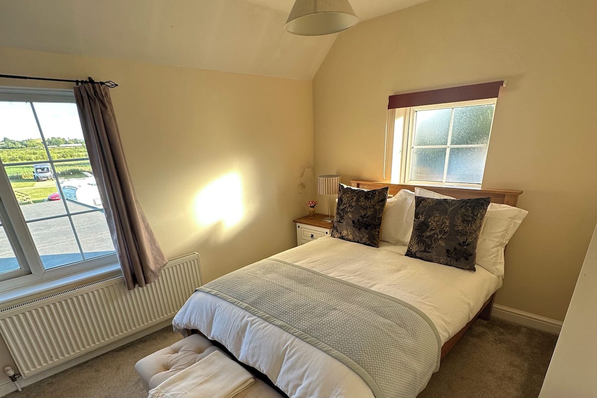 A bedroom with a large bed, a bench, a window with curtains, a radiator, and a ceiling light.