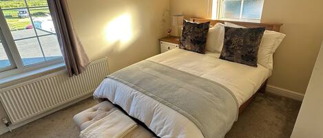A bedroom with a large bed, a bench, a window with curtains, a radiator, and a ceiling light.