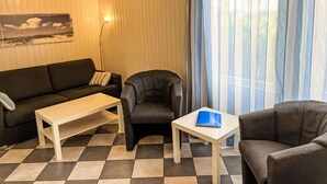 Ein gemütliches Zimmer mit kariertem Boden, einer Couch, einem Sessel, einem kleinen Tisch und einem Fenster mit Vorhängen.