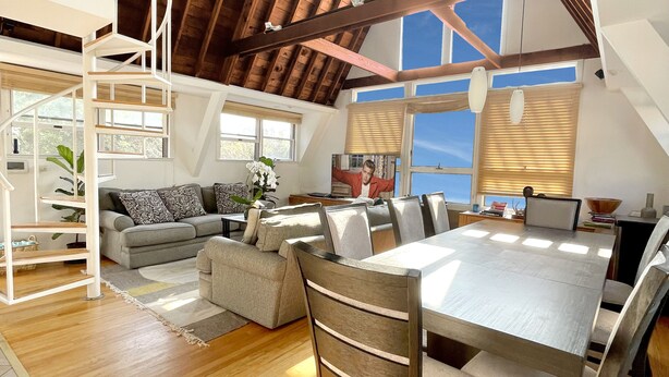 A spacious loft with a high ceiling, wooden beams, a dining area with a large table and chairs, a cozy seating area with a sofa, and a staircase leading to an upper level. - Immaculate Luxury Fire Island Pines Home, Gourmet Kitchen, Pool, Hot Tub Private (Fire Island Pines)