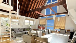 A spacious loft with a high ceiling, exposed wooden beams, a dining area with a large table and chairs, a cozy seating area with a sofa, and a staircase leading to an upper level. - Immaculate Luxury Fire Island Pines Home, Gourmet Kitchen, Pool, Hot Tub Private (Fire Island Pines)