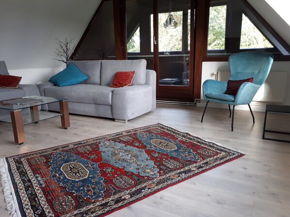 Ein modernes Wohnzimmer mit einem grauen Sofa, einem blauen Sessel, einem Glastisch und einem großen Teppich mit Muster.
