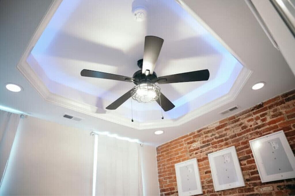 Ein Zimmer mit Deckenventilator, eingelassener Beleuchtung, einer Ziegelwand und einem Fenster mit weißen Vorhängen.