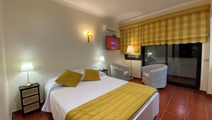 A hotel room with a bed, two bedside lamps, a TV, a window with a checkered curtain, and a chair.