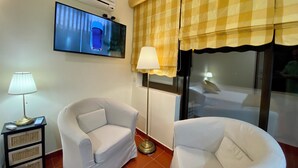 A hotel room with two white armchairs, a flat-screen TV mounted on the wall, an air conditioning unit, a checkered window valance, and a floor lamp.