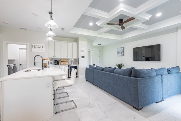 A modern kitchen with white cabinetry, a central island with bar stools, and a blue sofa in the living area. - Brand New Home near Warm Mineral Springs (North Port)