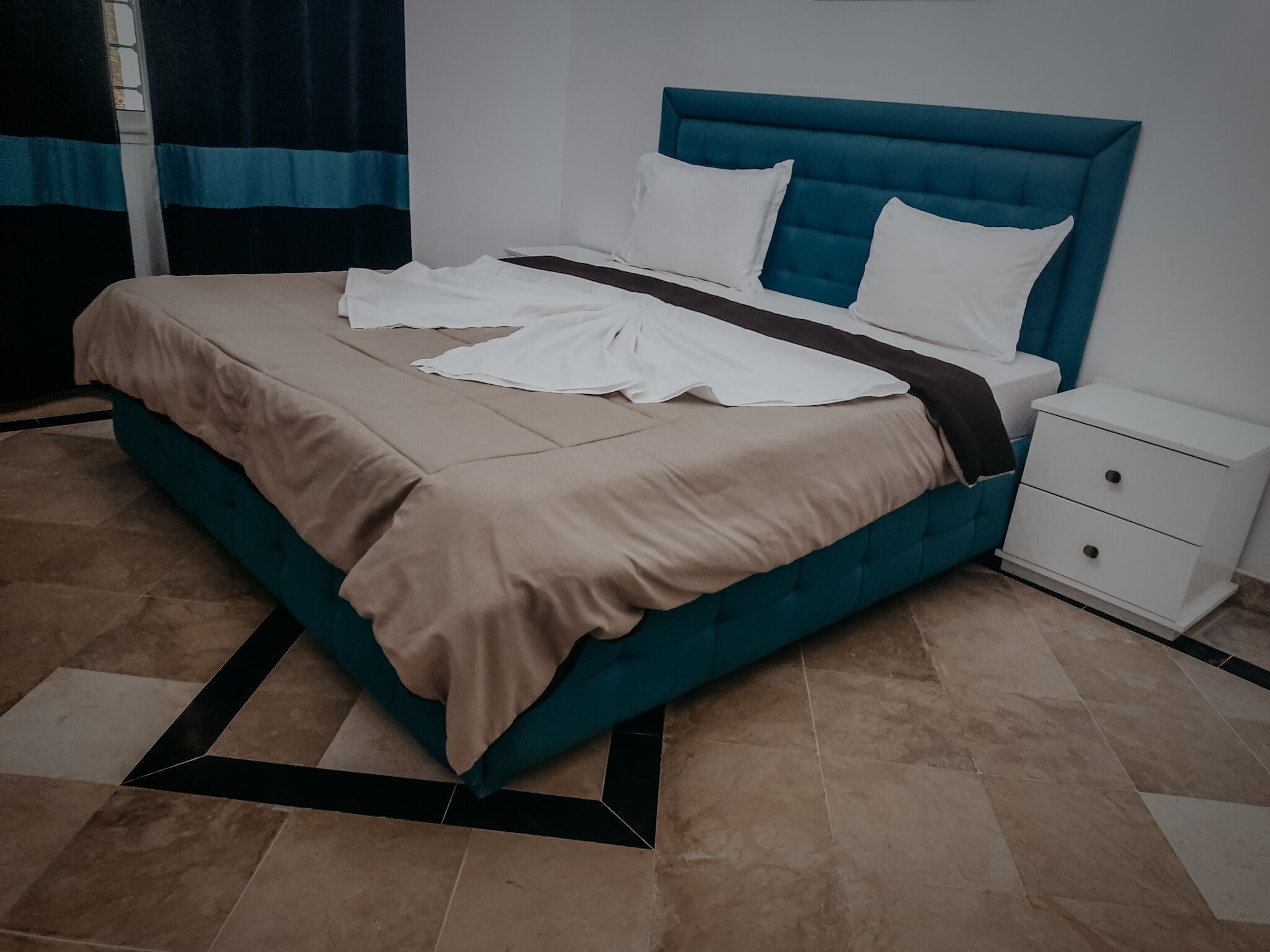 A neatly made bed with a teal headboard and white bedding, a white bedside table with drawers, and a patterned floor.