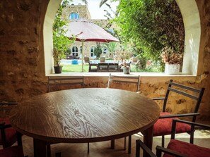 A round wooden table with chairs in a stone-walled room, overlooking a pool and outdoor seating area with a red umbrella. - Dar Mhenna de fêtes & séminaires (Hammamet)