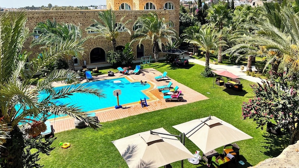 Un área de piscina con sillas de descanso y sombrillas, rodeada de palmeras y un edificio de piedra con ventanas de arco.