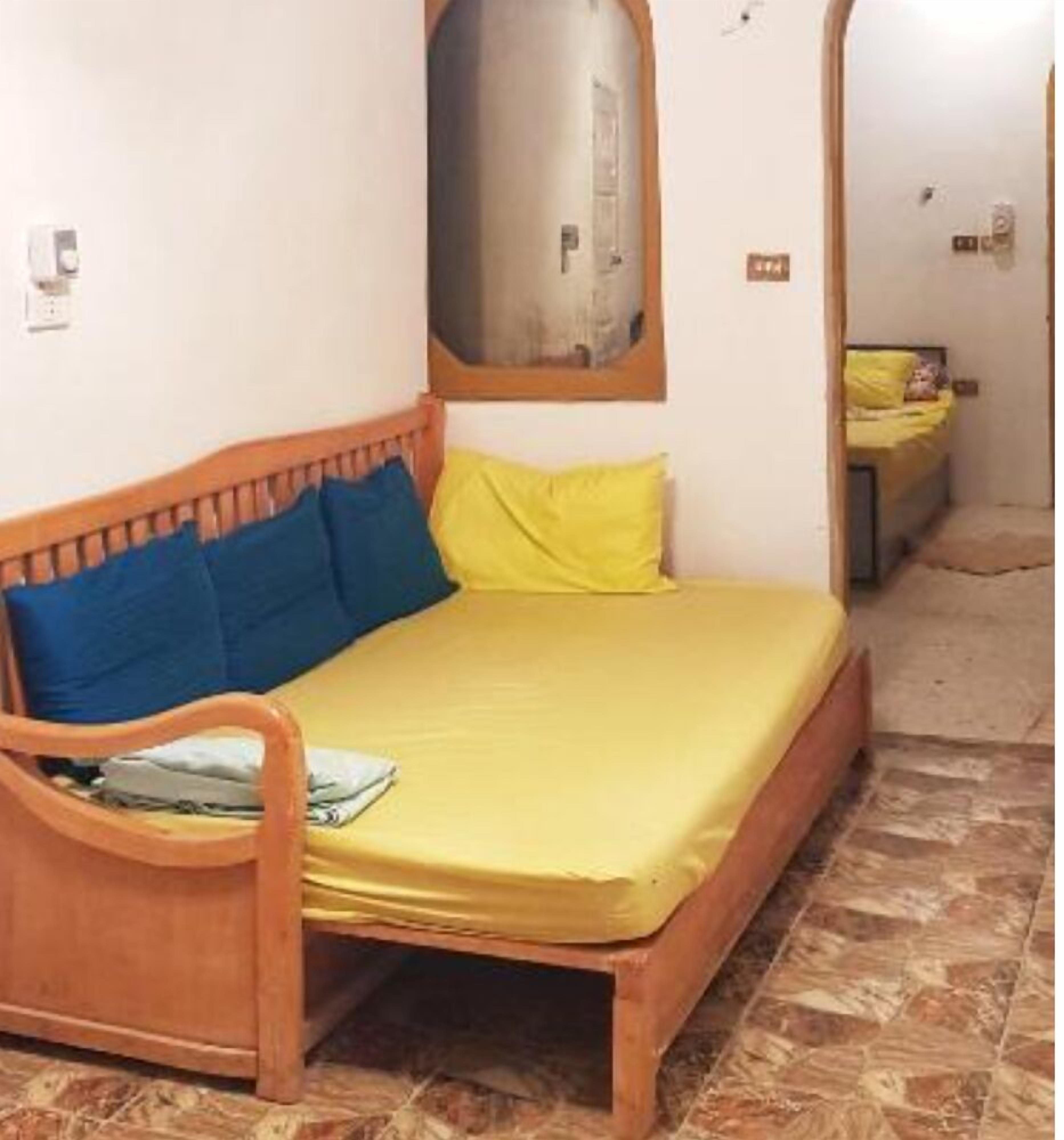 A single bed with a wooden frame and a yellow mattress. There is a window above the bed, and the room has a tiled floor.