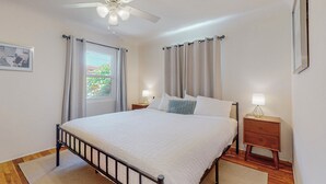 Ein Schlafzimmer mit Bett, Nachttisch, Deckenventilator und einem Fenster mit Vorhängen.