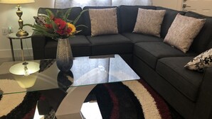 Una sala de estar con un sofá seccional gris oscuro, una mesa de centro de vidrio, un jarrón con flores, una lámpara y una alfombra estampada.