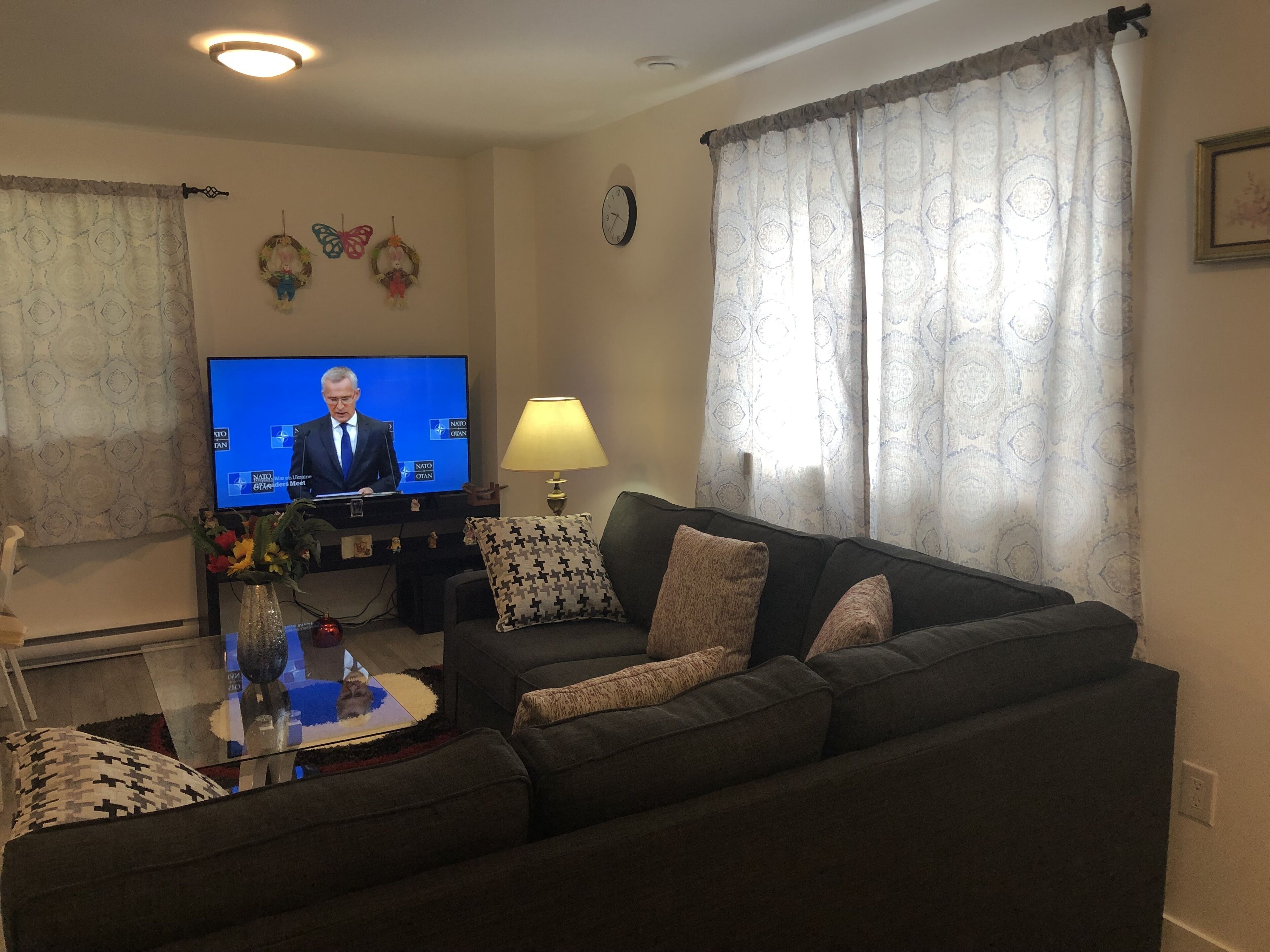 Ett vardagsrum med en soffa i mörk färg, ett soffbord i glas, en tv som visar ett nyhetsprogram, en väggklocka och mönstrade gardiner.
