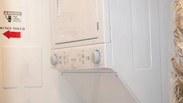Un appareil combiné lave-linge et sèche-linge blanc, de type à chargement vertical, muni d'un panneau d'avertissement indiquant « Ne pas toucher » et d'un avertissement indiquant « Haute tension ».