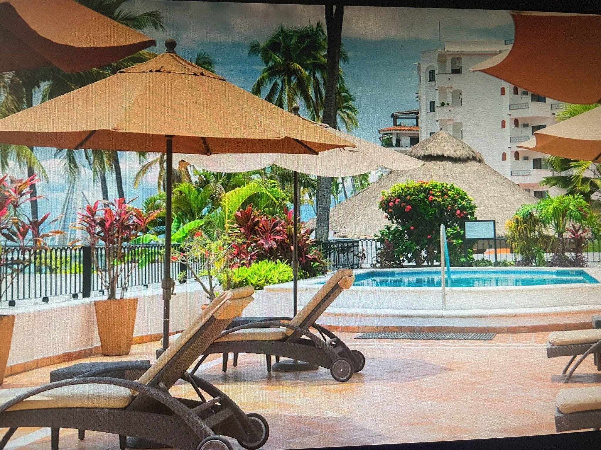 A poolside area with lounge chairs and umbrellas, surrounded by tropical plants and a thatched-roof structure.