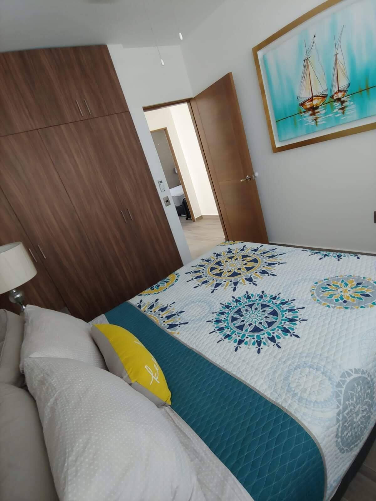 A bedroom with a bed, a wooden wardrobe, a painting of sailboats, and a door leading to another room.