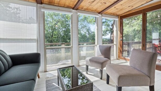 A sunlit room with a wooden ceiling, large windows, a grey sofa, a beige armchair, and a glass coffee table. - Lake House Landing w/ view of sailboat cove, quiet street (Garfield)