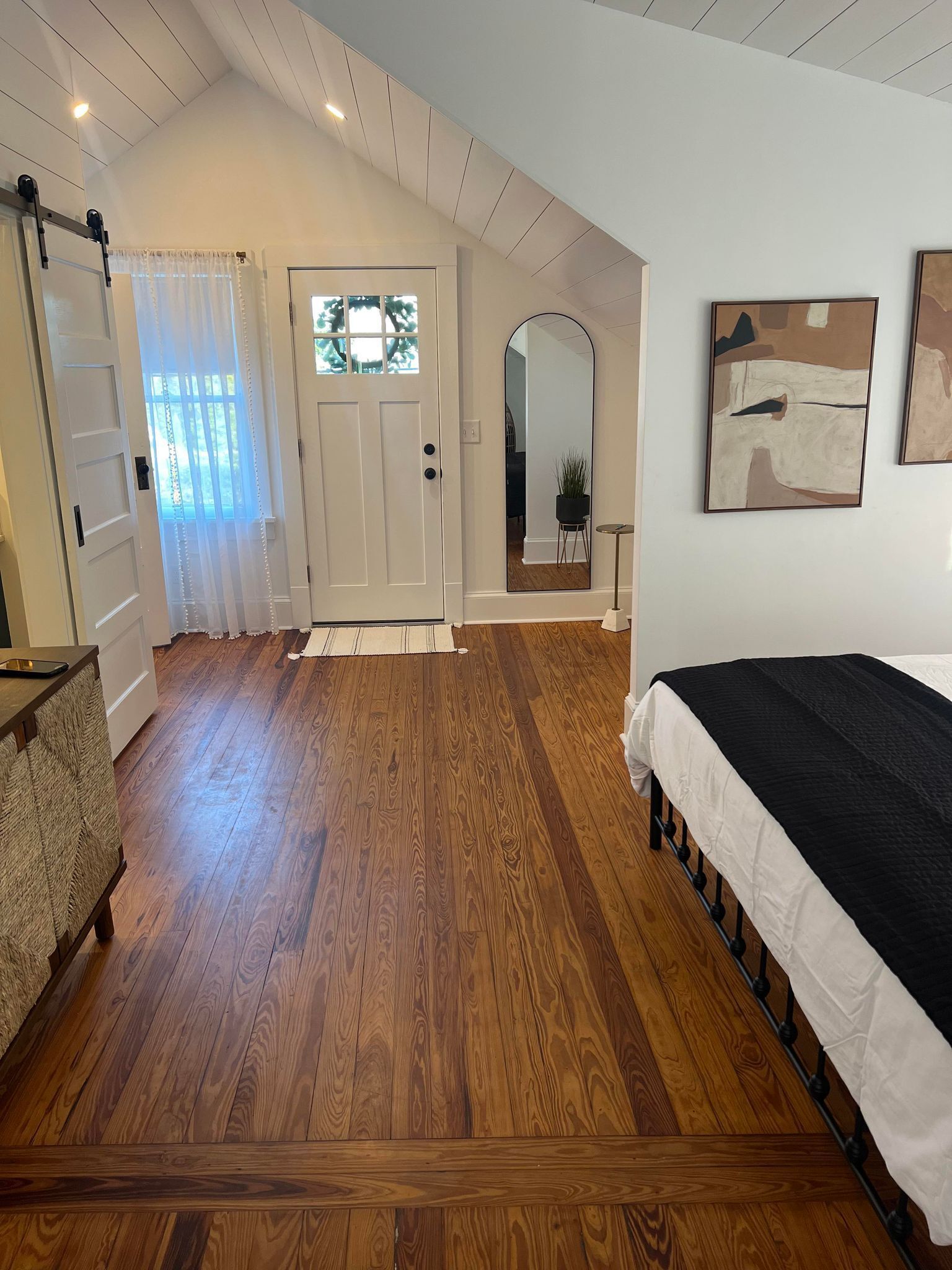Une pièce avec un plancher en bois, un lit, une porte, un miroir et un tableau accroché au mur.