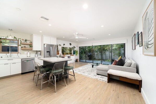 A modern kitchen with white cabinets, a dining area with a table and chairs, and a large window offering a view of greenery.