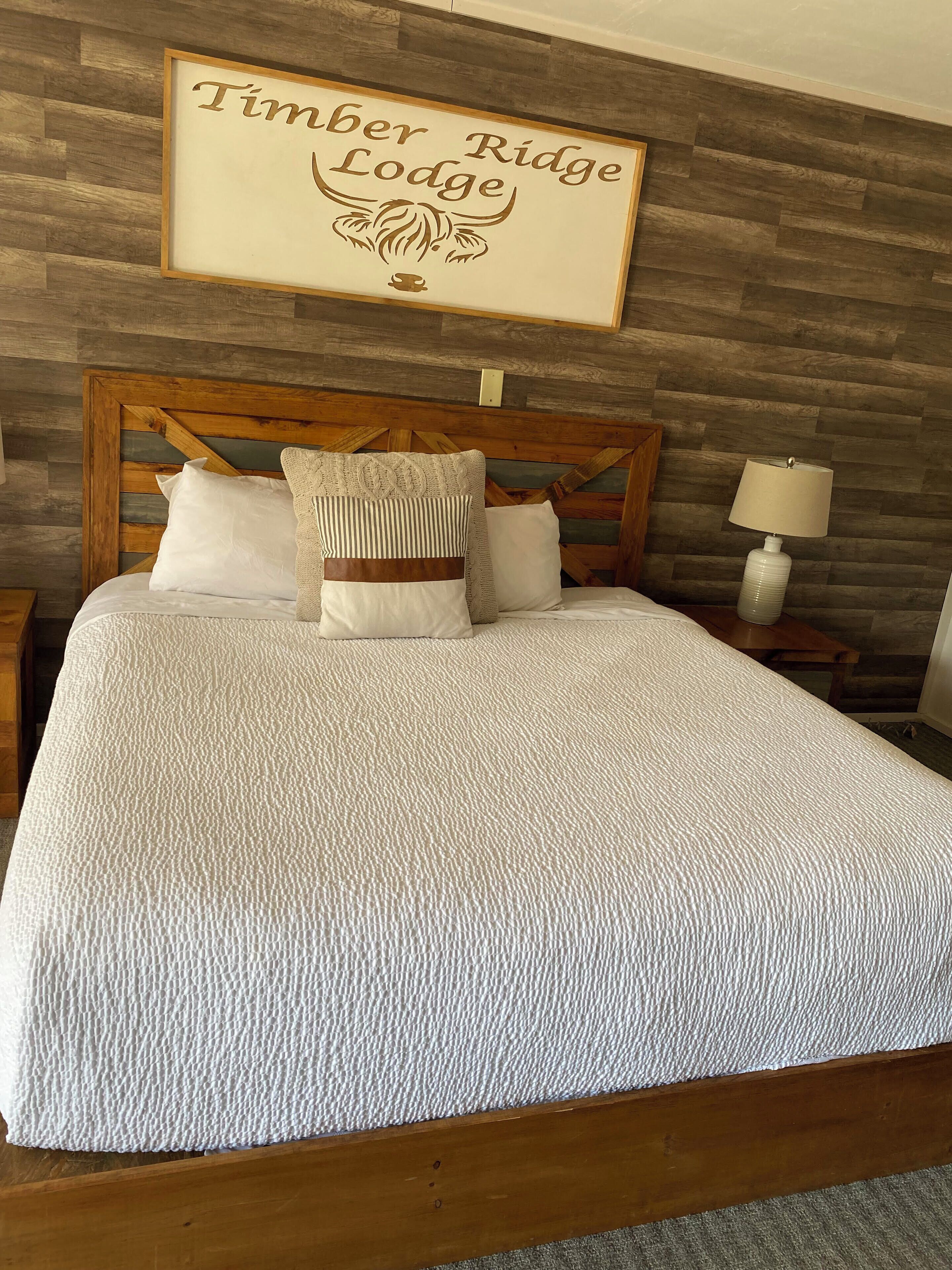 Una cama bien hecha con una colcha blanca texturizada, un cabecero de madera y un cartel que dice 'Timber Ridge Lodge'.