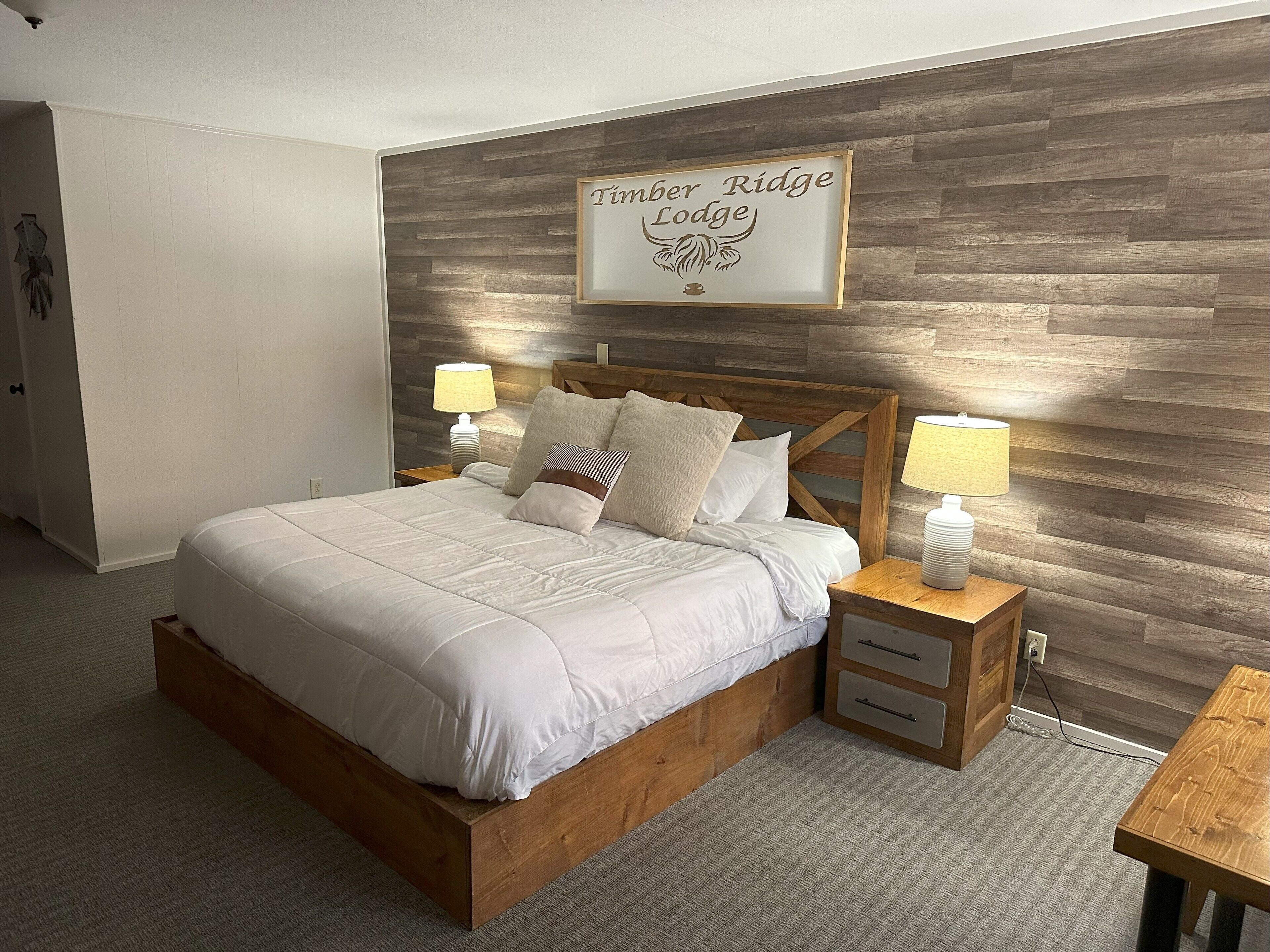 En pænt redt seng med et sengegavl, to sengeborde med lamper, og et skilt over sengen med teksten 'Timber Ridge Lodge'.