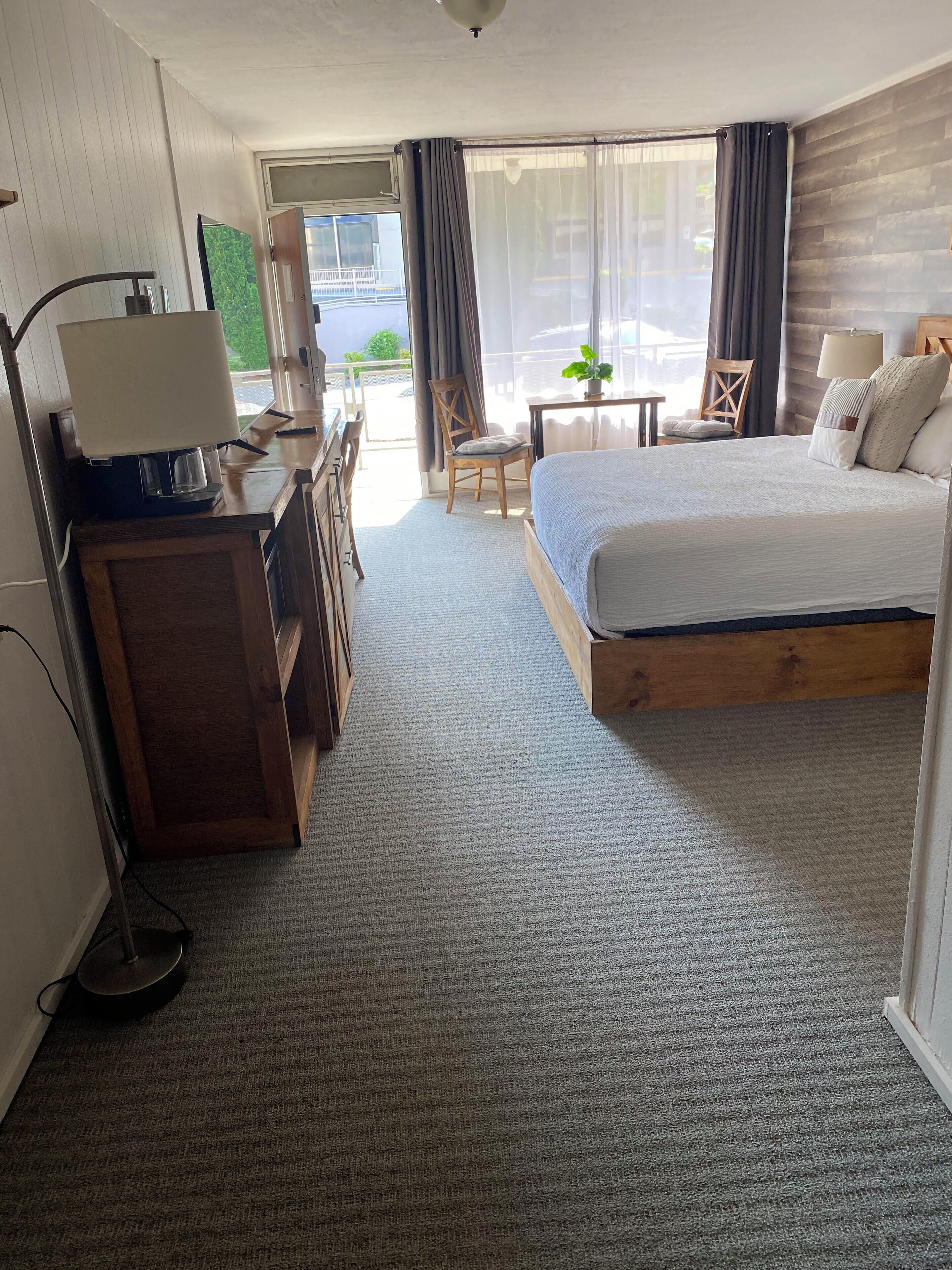 Habitación de hotel con cama, escritorio con lámpara, una silla, una mesita con planta y vistas al exterior.
