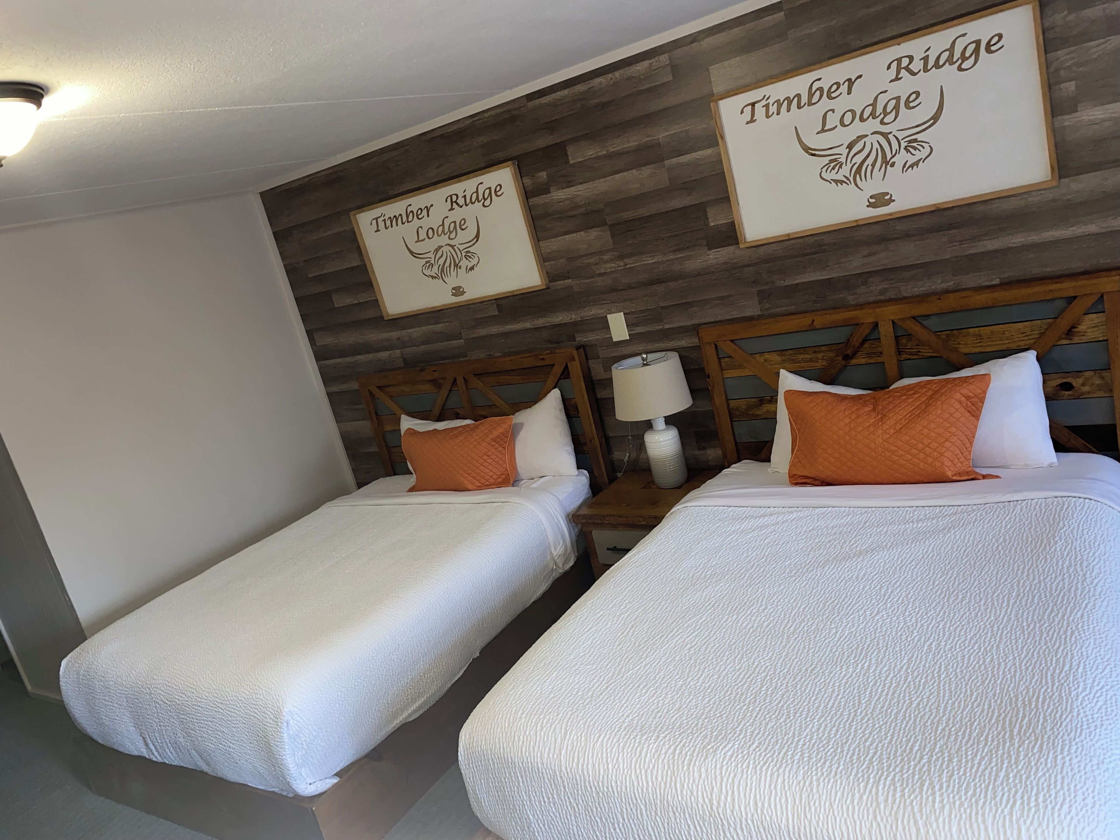 Twee enkele bedden met witte beddengoed en oranje kussens in een kamer met houten paneelwanden. Op de muur staat een bordje met de tekst 'Timber Ridge Lodge'.