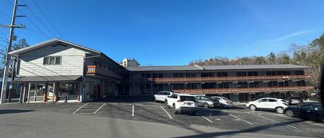 Ein Parkplatz mit mehreren Autos, ein Gebäude mit flachem Dach und vielen Fenstern, und ein klarer, blauer Himmel.