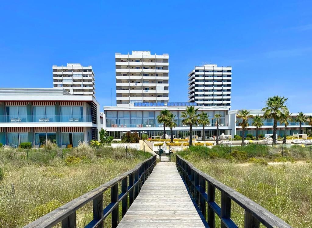 A wooden boardwalk leading to a modern building complex with multiple high-rise structures in the background.