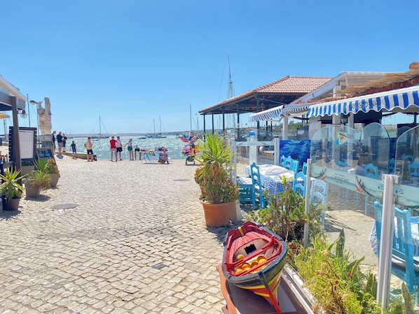 A cobblestone walkway leading to a waterfront area with boats, outdoor seating, and a clear view of the sea.