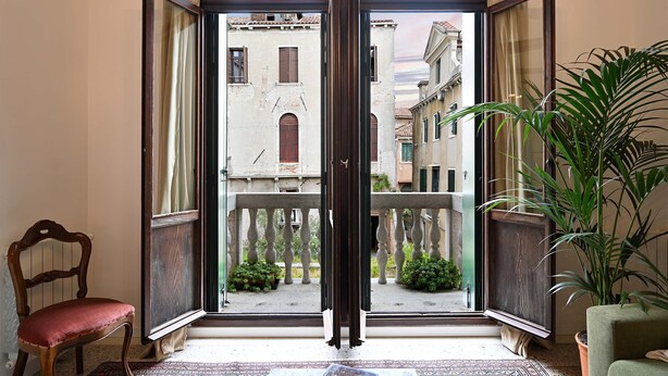 Ein Zimmer mit einer großen Glasschiebetür, die zu einem Innenhof mit Blick auf historische Gebäude führt. Im Zimmer steht ein Sessel und ein Blumentopf.