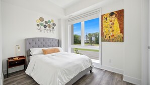 A bedroom with a bed, a nightstand, a wall art piece, and a window. - Exquisite, bright &modern 2 bedroom condo/Free Parking (Niagara Falls)