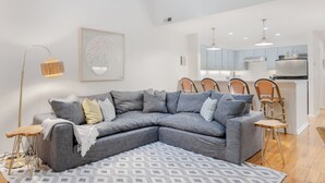 A modern living room with a grey sectional sofa, a patterned rug, and a dining area with wicker chairs.