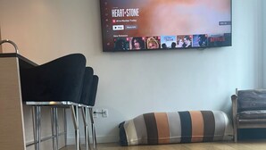 Ett modernt vardagsrum med trägolv, en svart soffa, en randig pall och en tv som är monterad på väggen och visar filmen ”Heart Stone”.