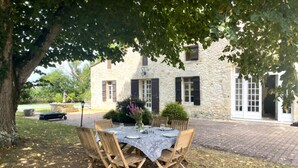 A stone building with a paved courtyard, a wooden table set for dining, and a large tree providing shade.