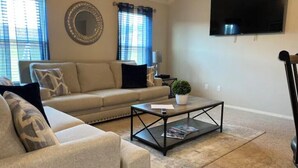 Un salon avec un canapé beige, une table basse, un téléviseur fixé au mur et une fenêtre avec des rideaux.