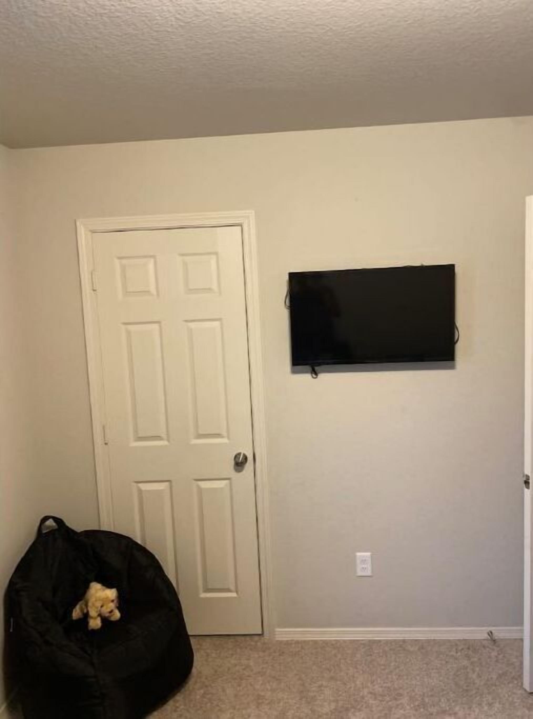 Una habitación con una puerta blanca, un televisor de pantalla plana montado en la pared y un sillón informal negro con un pequeño peluche.