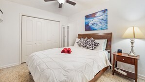 A bedroom with a bed, a ceiling fan, a nightstand with a lamp, and a painting on the wall.