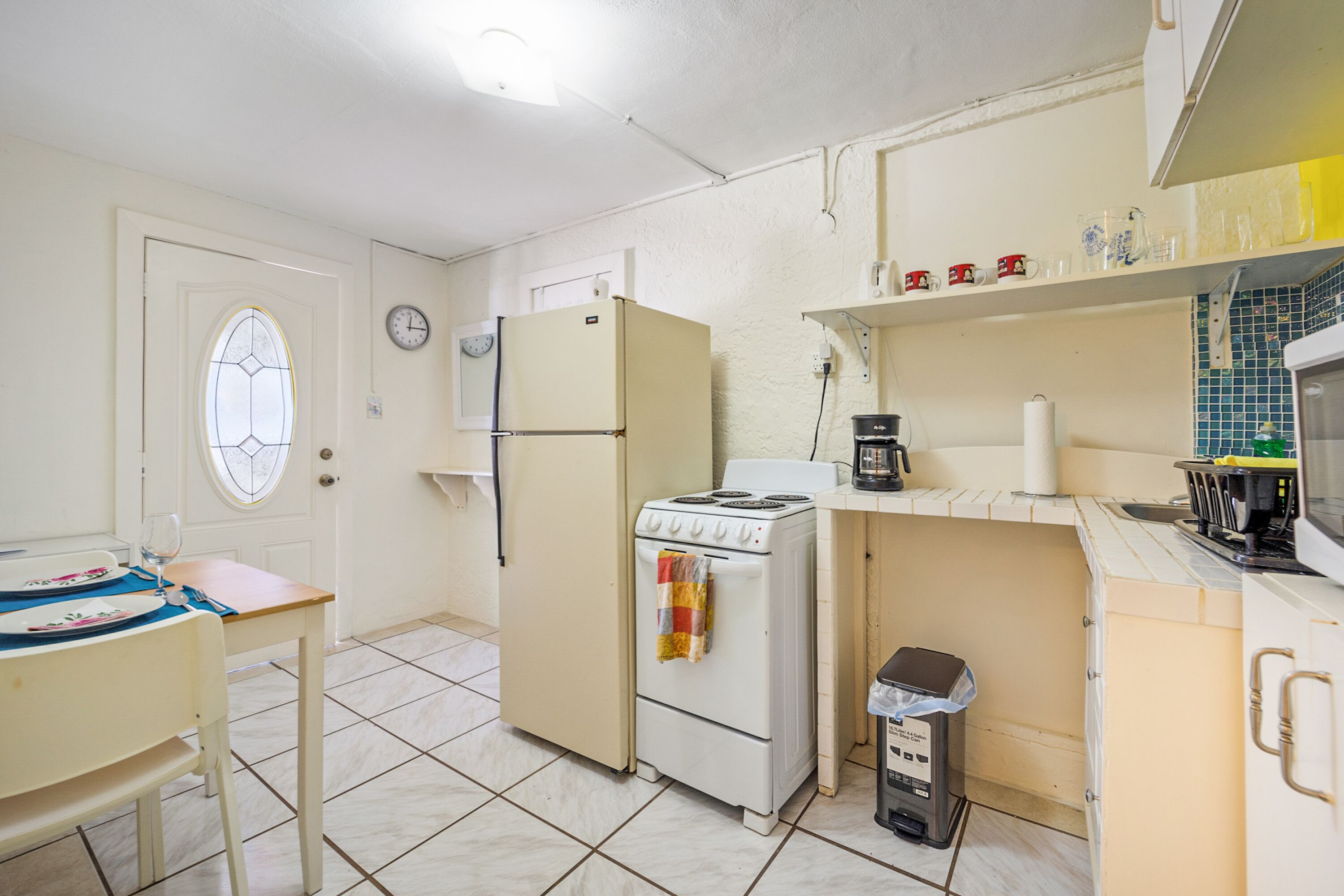 A compact kitchen with a refrigerator, stove, and microwave. There is a small trash bin and a window with a decorative glass design.