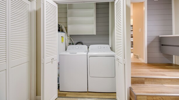 Ein Waschbereich mit zwei weißen, frontgeladenen Waschmaschinen und Trocknern, einer kleinen Ablage für Reinigungsmittel und einer Holztreppe, die in eine obere Ebene führt.