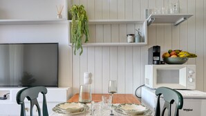 Spiseområde med et træbord, hvide stole, et fjernsyn, en mikrobølgeovn og en skål med frugt på en hylde.