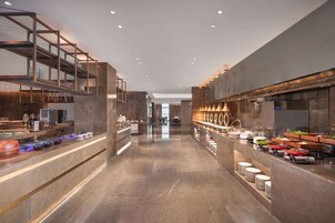 Un moderno vestíbulo de hotel con una escalera central, una zona de buffet y un área de descanso con mesas y sillas.