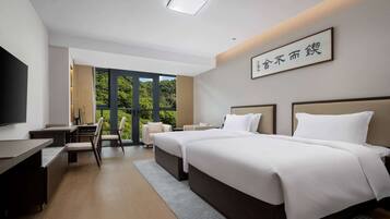 A modern hotel room with a large bed, a desk, and a TV. There is a window with a view of greenery outside.