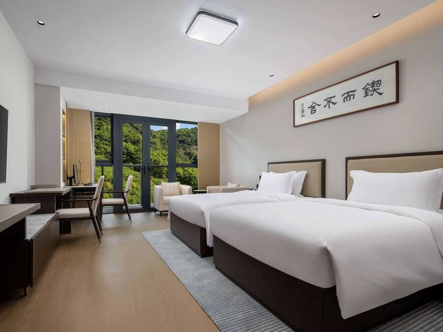A modern hotel room with a large bed, a desk, and a TV. There is a window with a view of greenery outside.